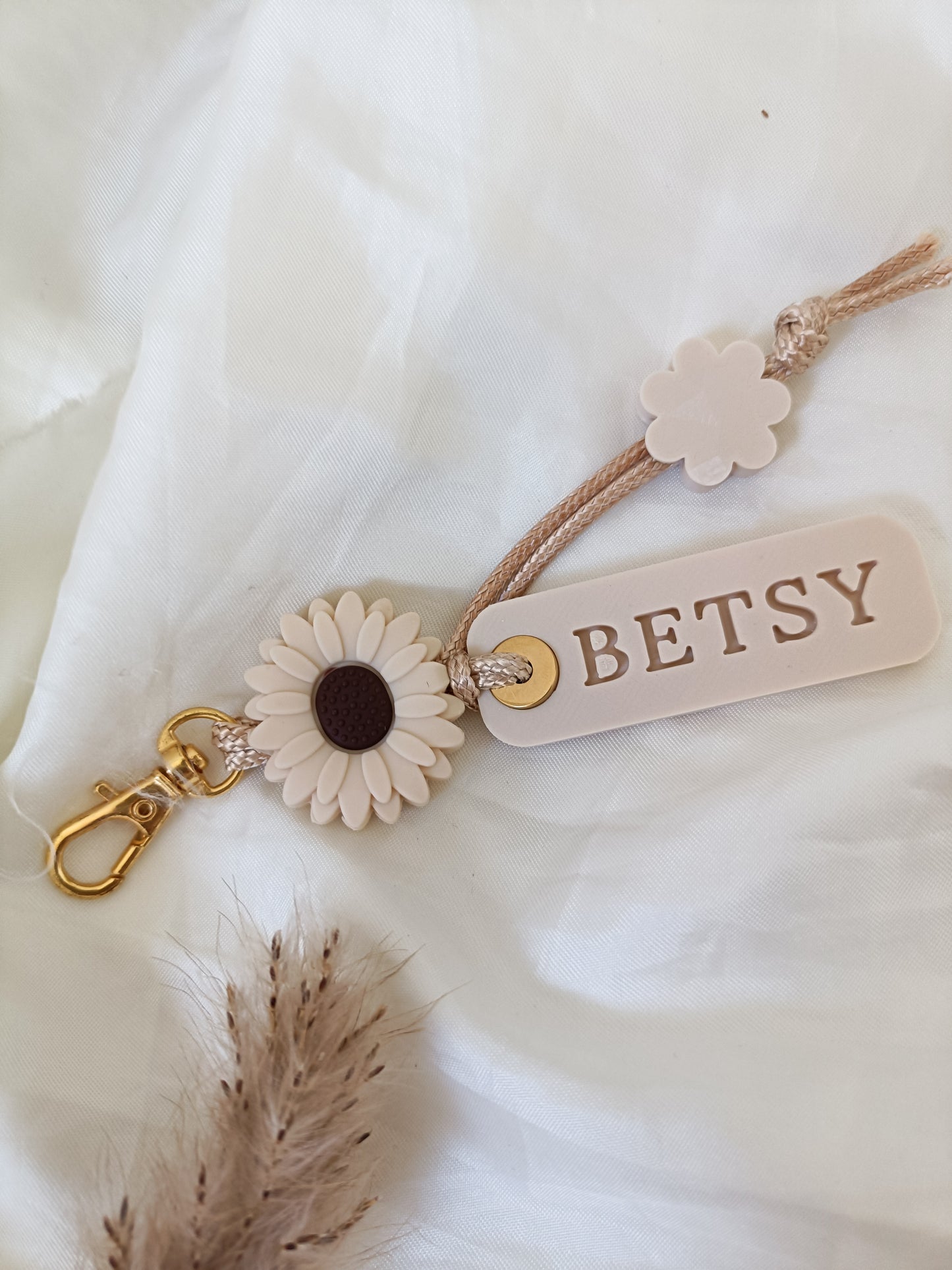 Custom name keyring with ivory daisy charm, gold lobster clasp, and braided beige cord, personalized tag reading "BETSY" on a white fabric background.