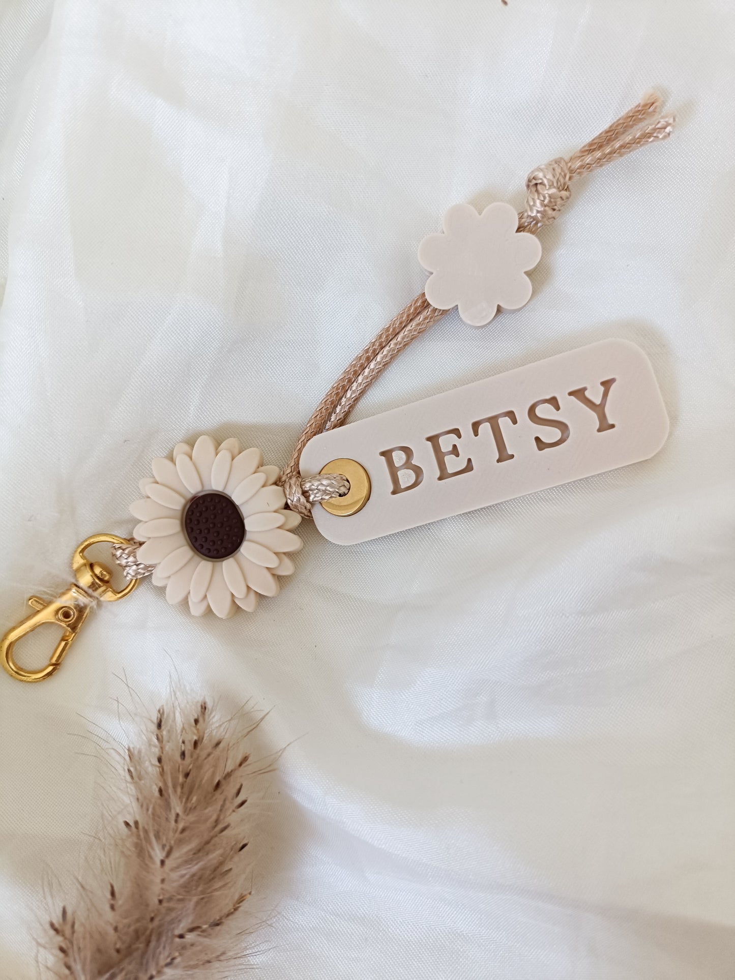 A personalized keychain or bag charm with a beige rope strap, a cream-colored acrylic tag engraved with 'BETSY', a cream flower accent, a faux sunflower with a dark brown center, and a gold-tone lobster clasp, displayed on a soft white fabric background.