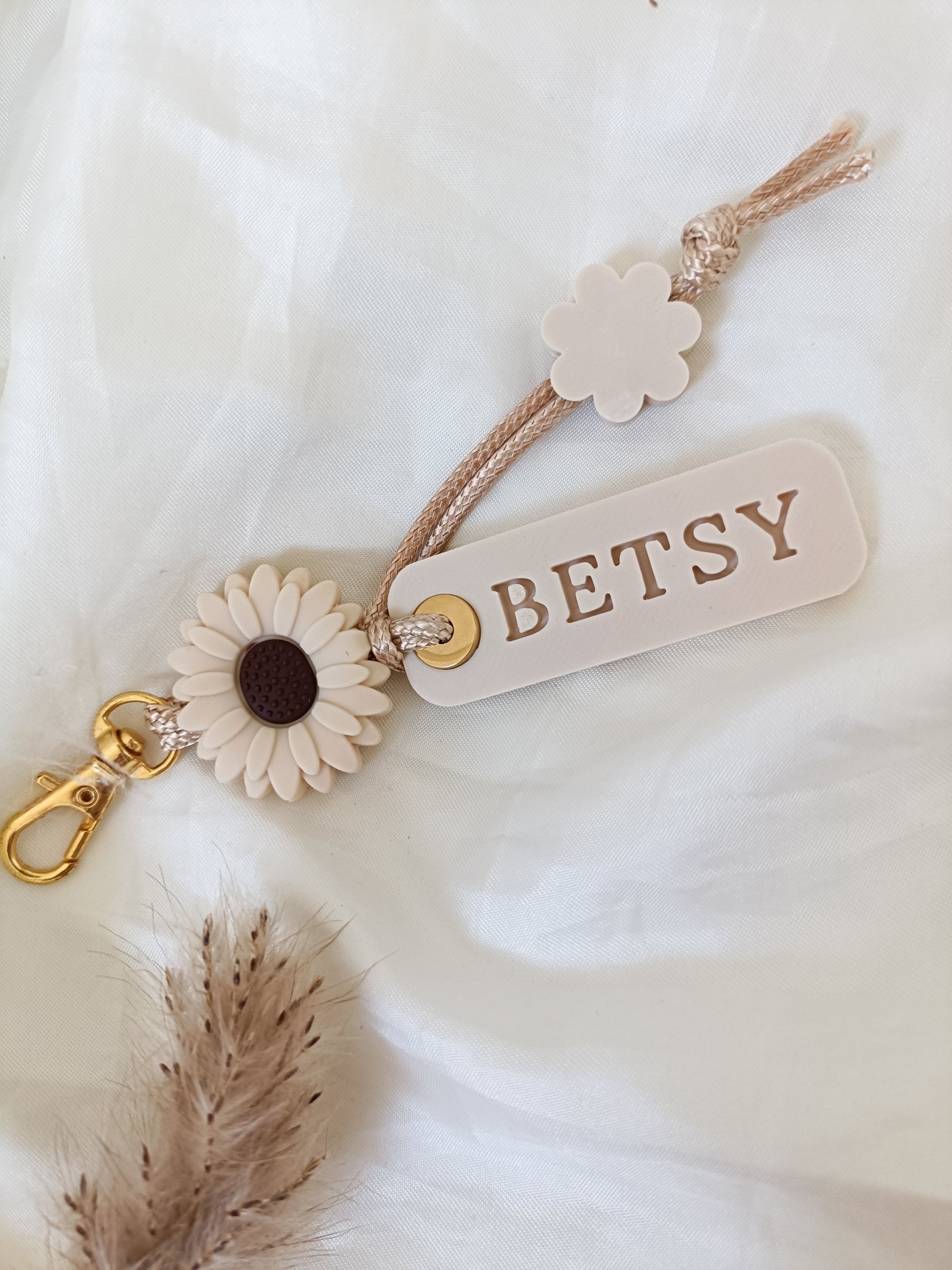 A personalized keychain or bag charm with a beige rope strap, a cream-colored acrylic tag engraved with 'BETSY', a cream flower accent, a faux sunflower with a dark brown center, and a gold-tone lobster clasp, displayed on a soft white fabric background.