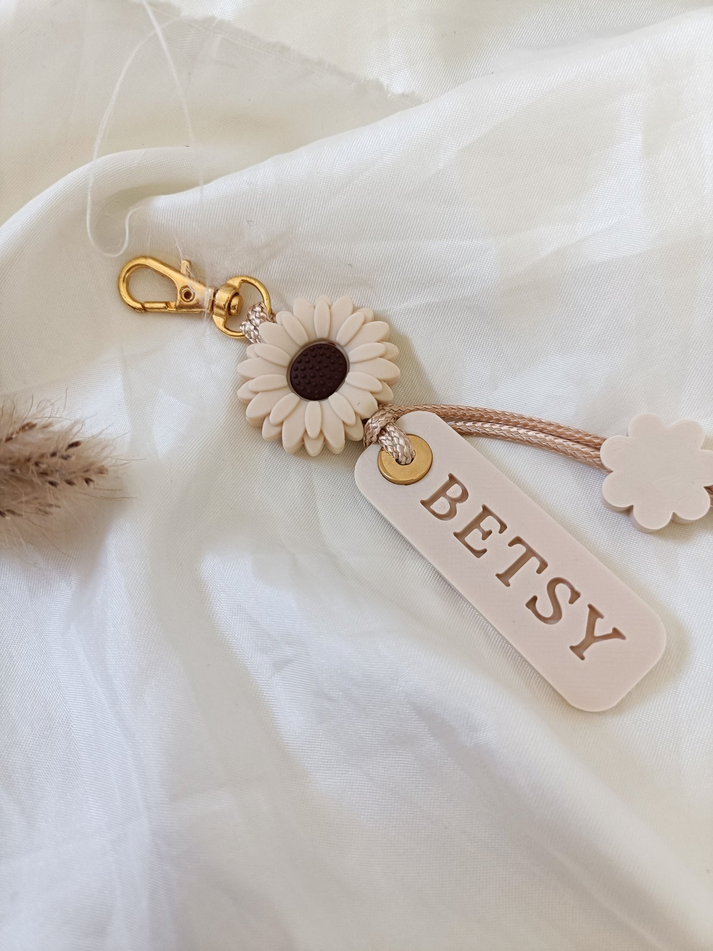 A beige and white sunflower keychain with a gold clasp, a hanging tag that reads 'BETSY', and a small flower charm, styled on a white fabric background.