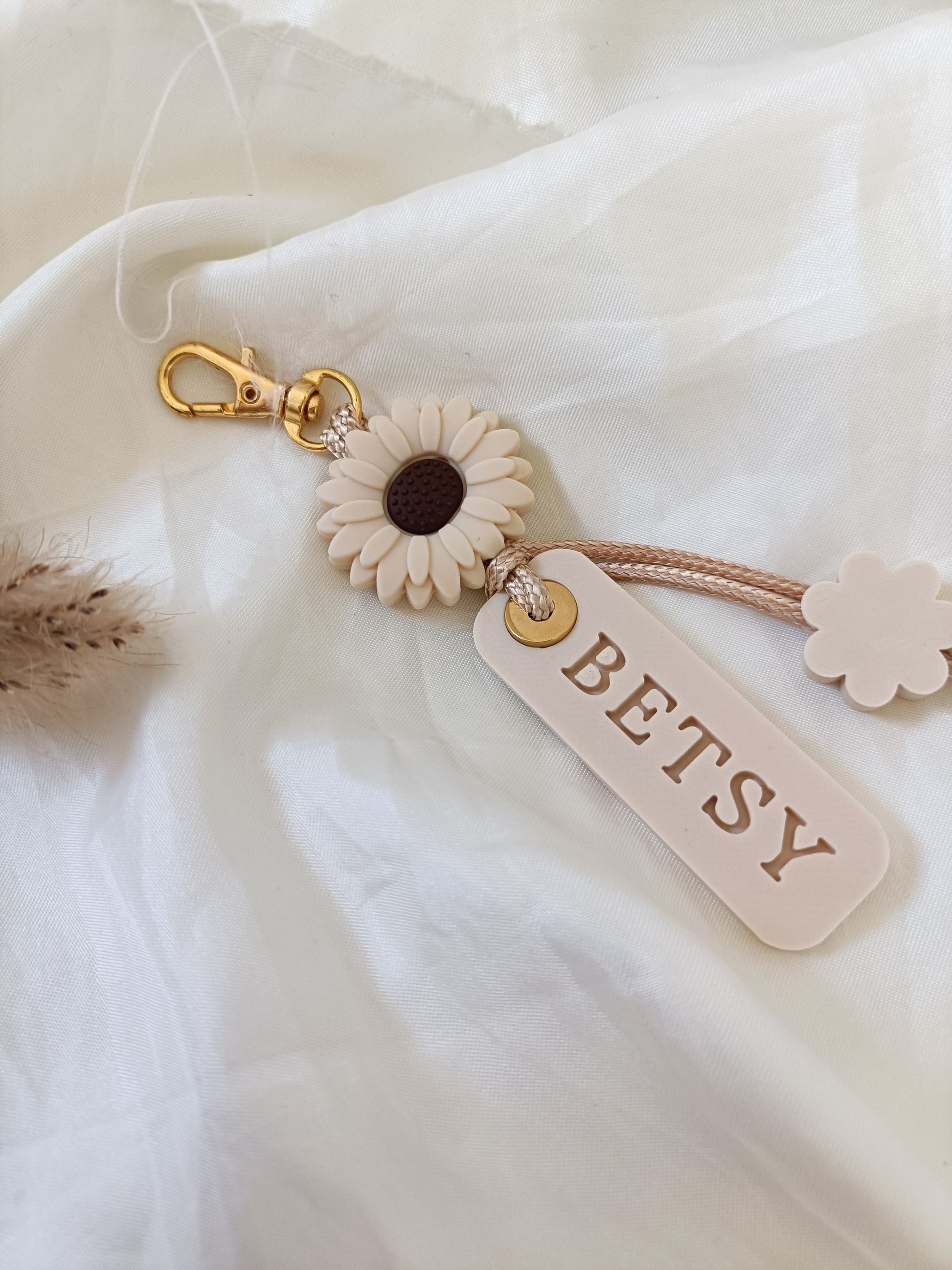 A beige and white sunflower keychain with a gold clasp, a hanging tag that reads 'BETSY', and a small flower charm, styled on a white fabric background.