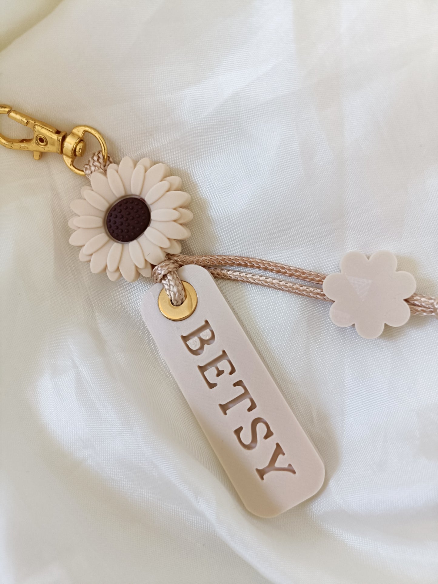 A personalized keychain featuring a cream-colored daisy with a dark brown center, a cream flower charm, and a rectangular cream tag engraved with the name 'BETSY'. The keychain is attached to a beige braided cord and a gold-tone lobster clasp, displayed on a soft white fabric background.