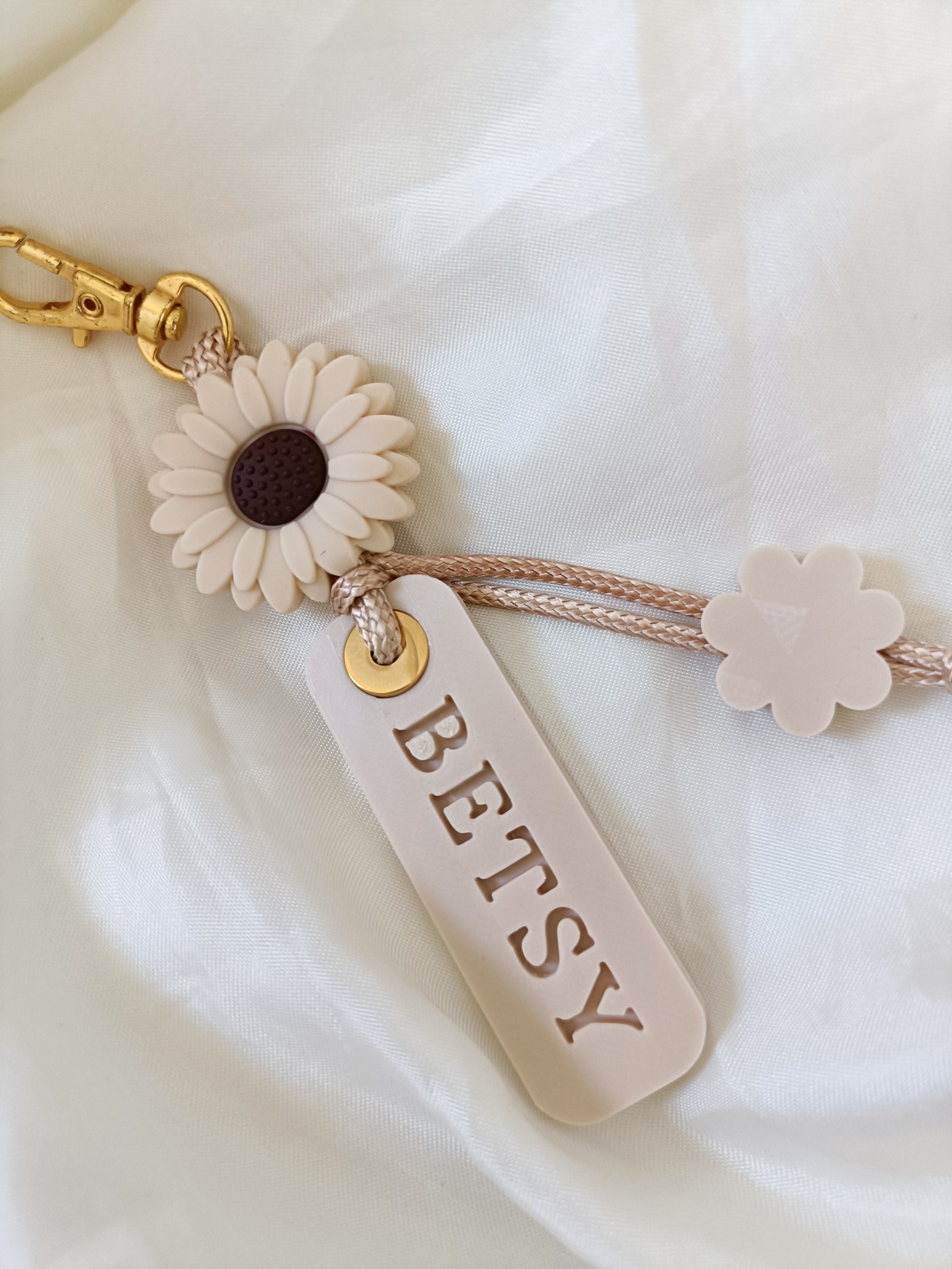 A personalized keychain featuring a cream-colored daisy with a dark brown center, a cream flower charm, and a rectangular cream tag engraved with the name 'BETSY'. The keychain is attached to a beige braided cord and a gold-tone lobster clasp, displayed on a soft white fabric background.