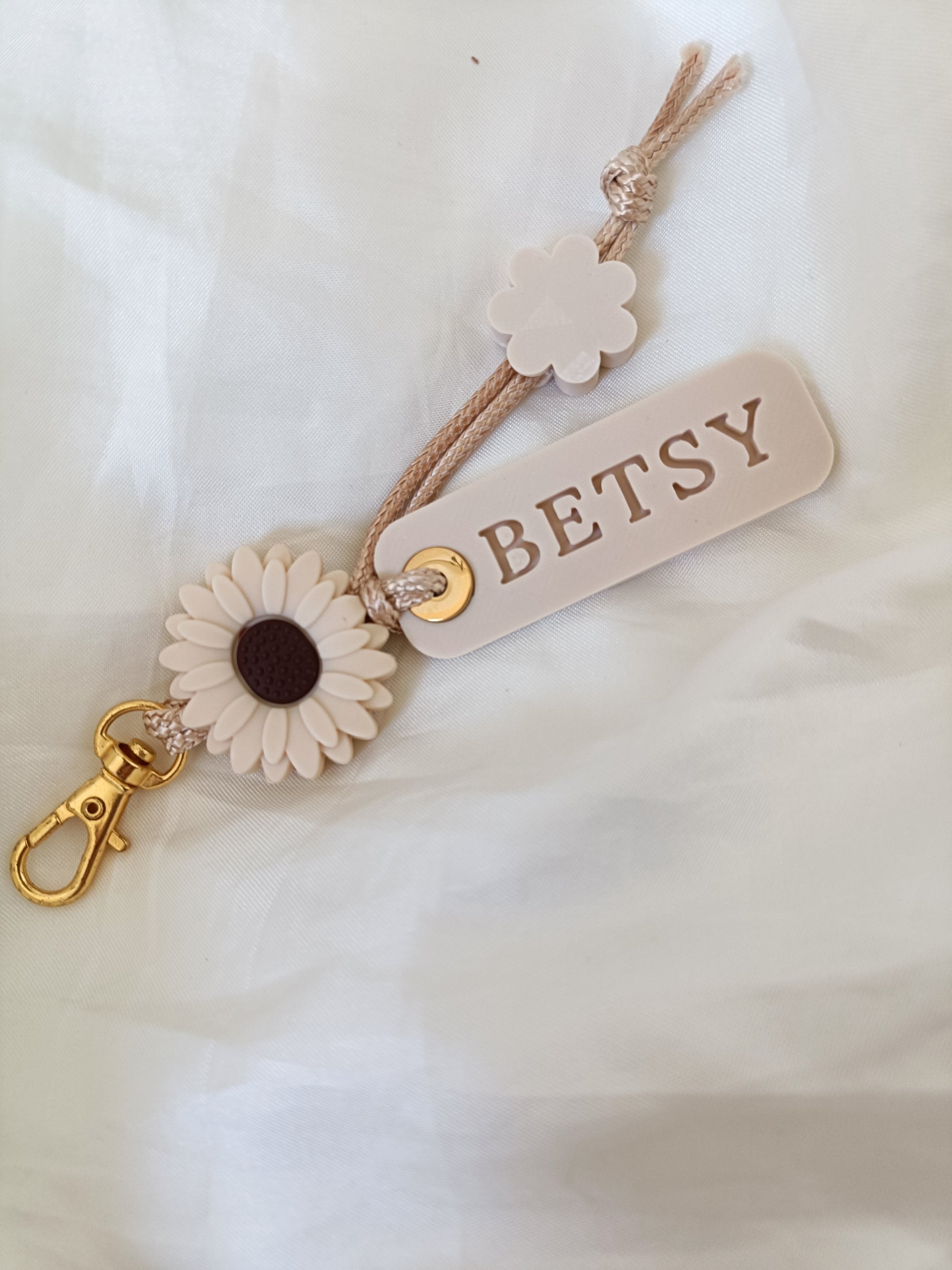 A personalized keychain or bag charm featuring a cream-colored faux leather tag with the name 'BETSY' in brown lettering, a cream flower charm, a brown cord, and a gold-tone lobster clasp, displayed on a soft white fabric background.